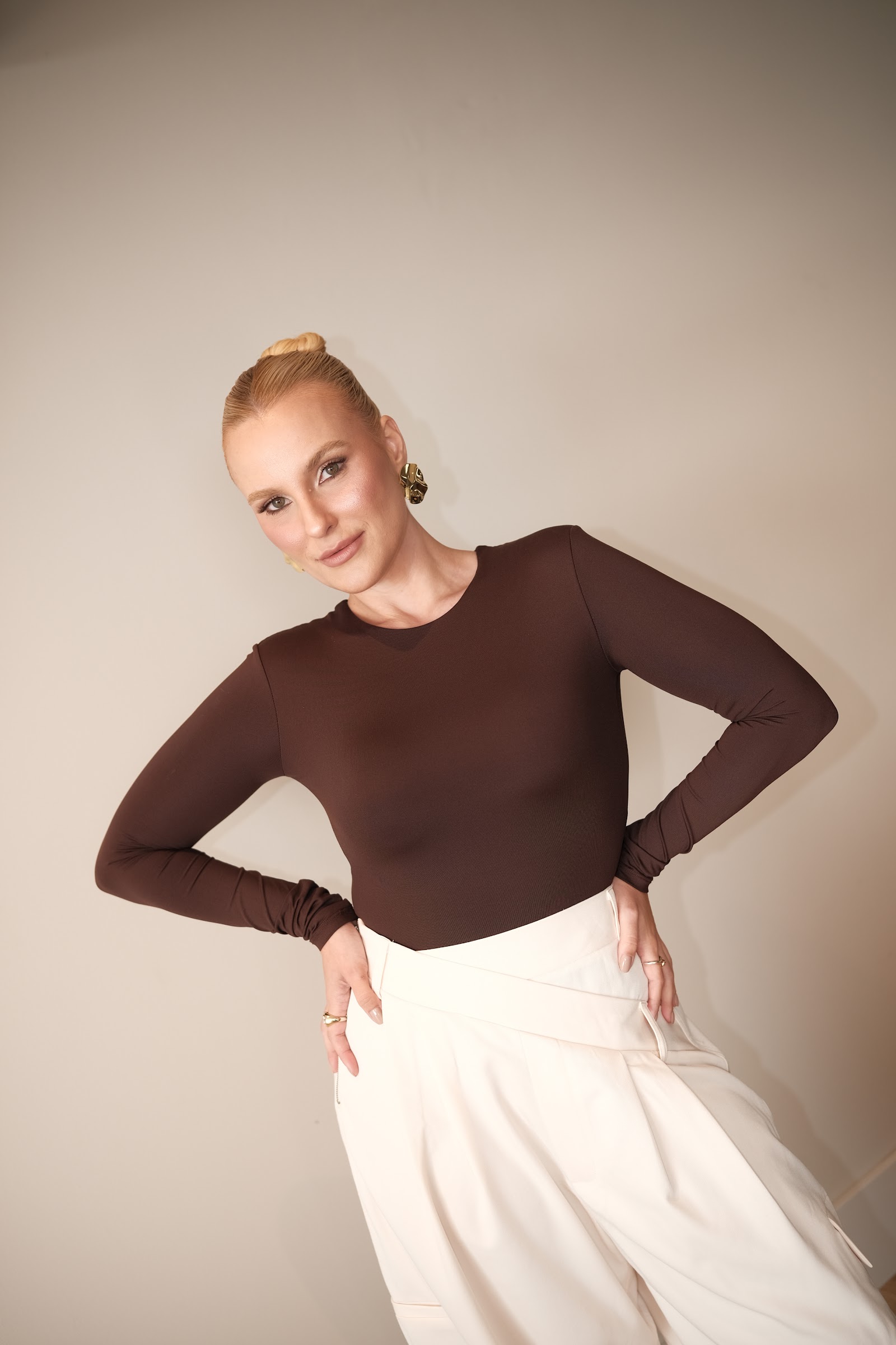 Gabriela Wiggers, de body marrom e calça off-white, olhando para a câmera com as mãos na cintura